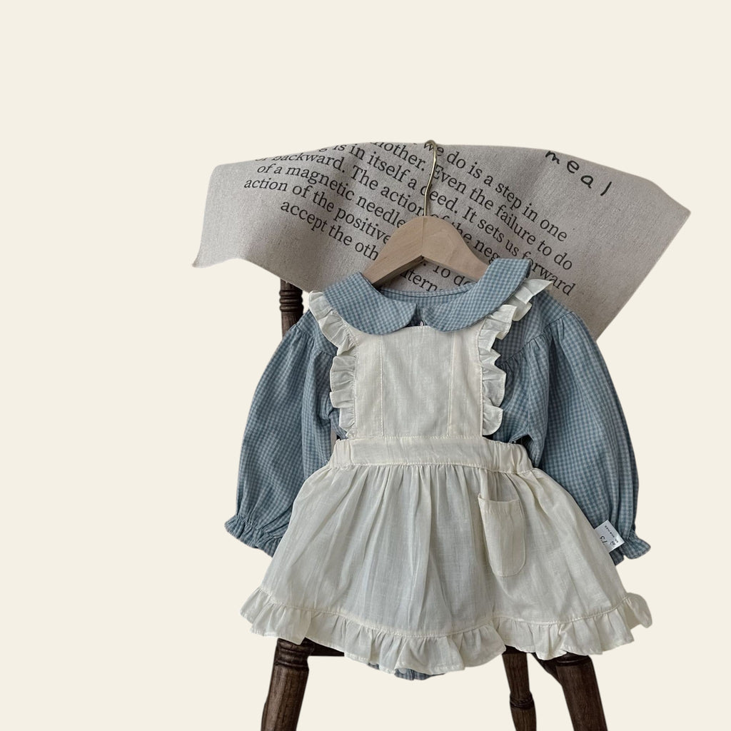 Children's outfit with white dress and blue checkered shirt on a wooden hanger against a beige background.