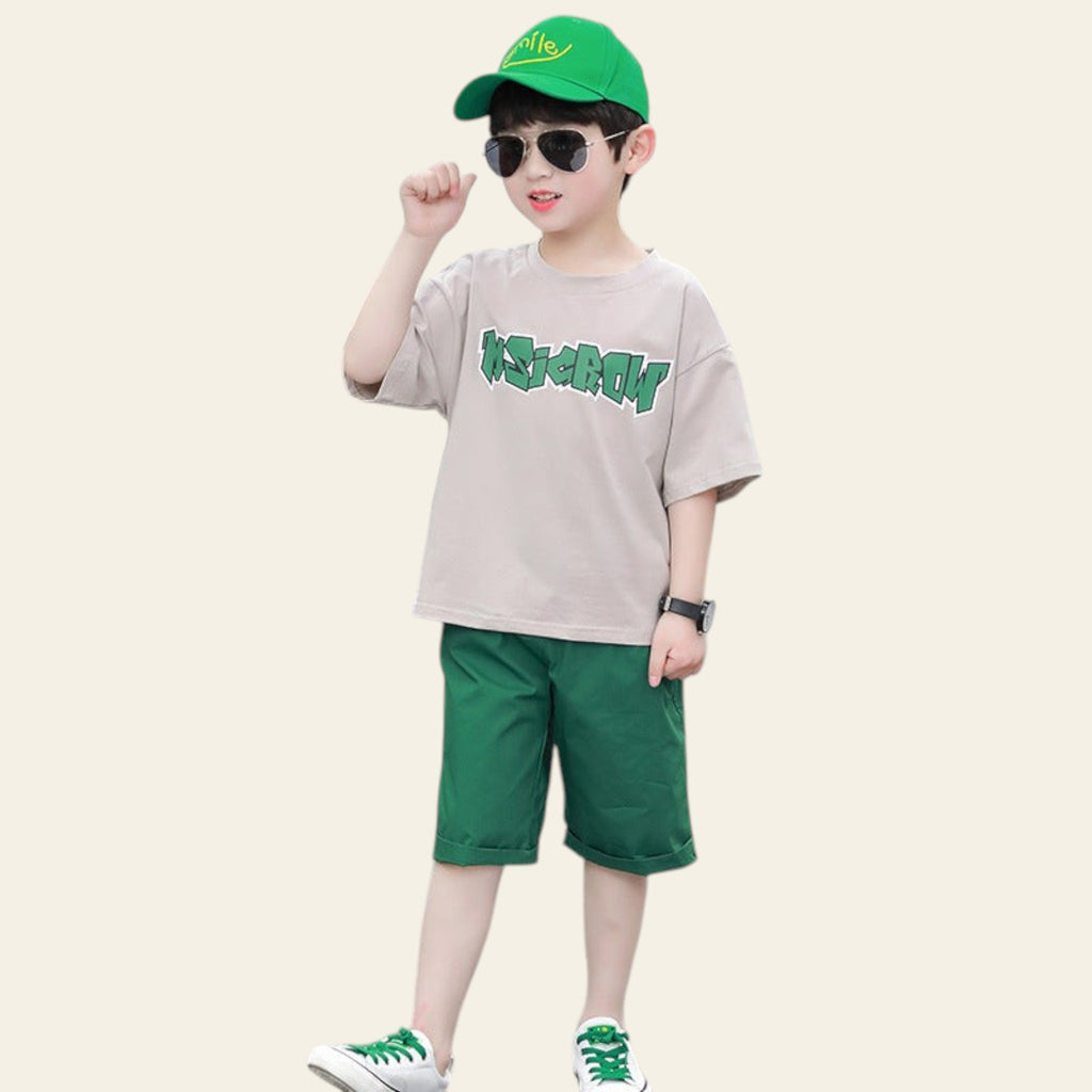 Child wearing a beige shirt with green text, green shorts, and a green cap on a street.