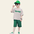 Child wearing a beige shirt with green text, green shorts, and a green cap on a street.
