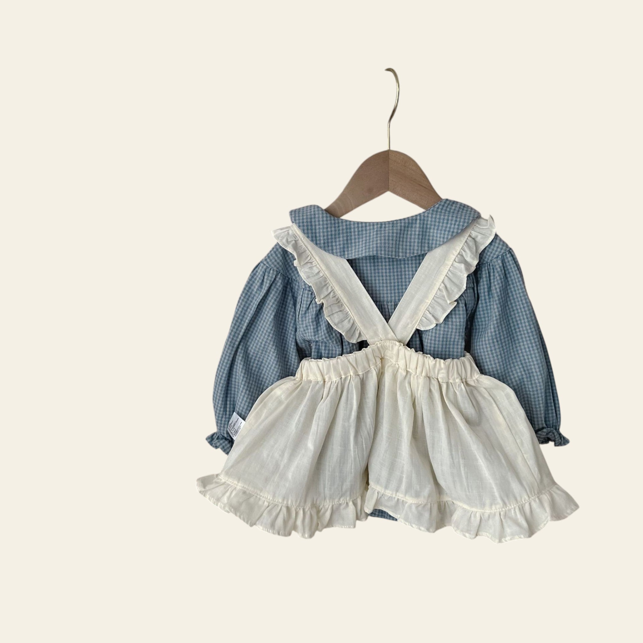 Children's outfit with blue checkered shirt and white ruffled shorts on a hanger against a neutral background.