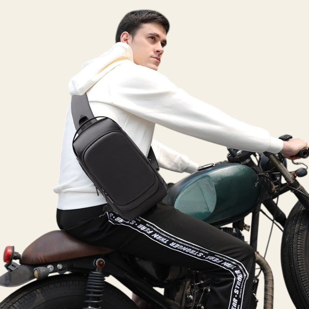 Man sitting on a motorcycle wearing a black crossbody bag against a gray wall.