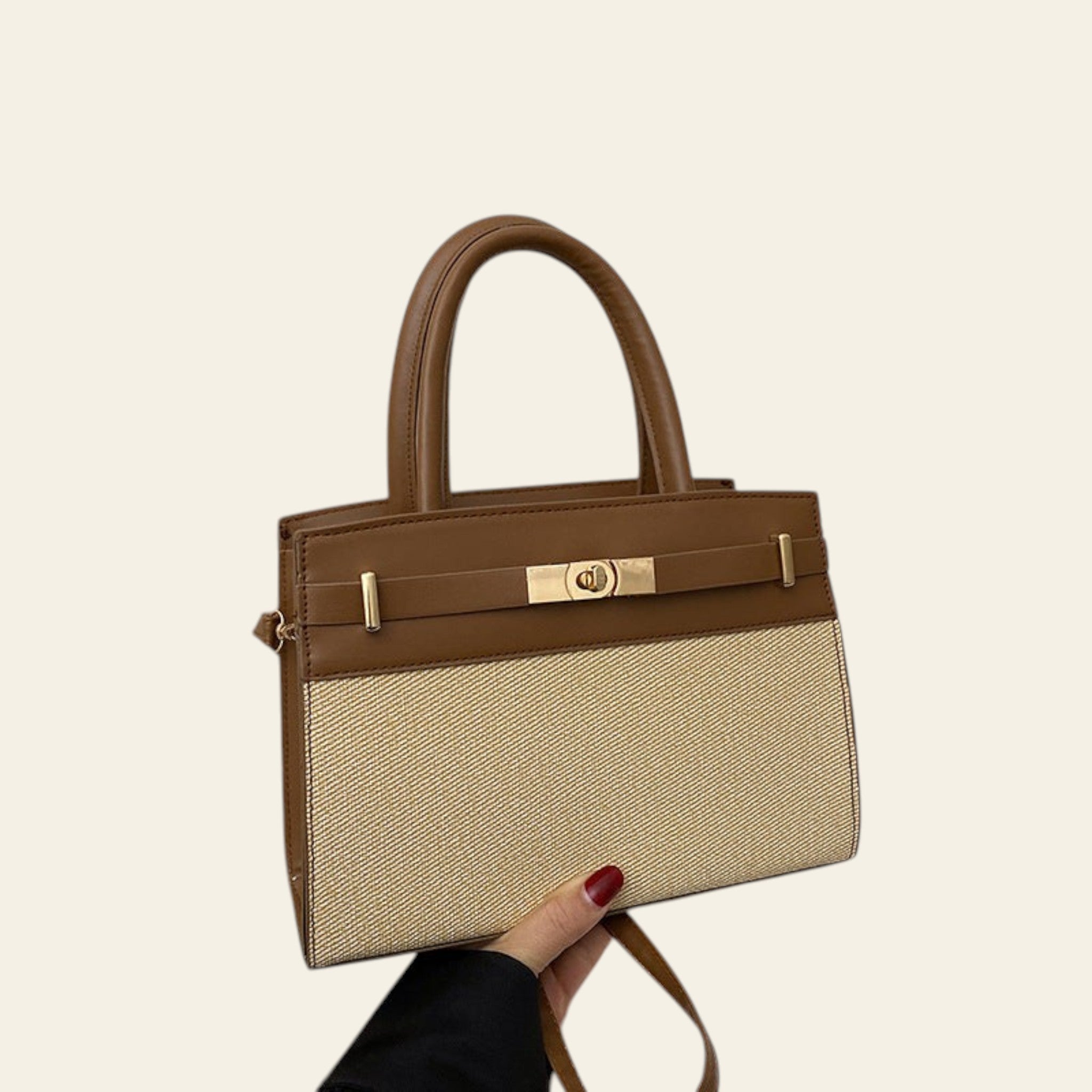 Brown and beige handbag held by a hand on a white surface with a beige jacket in the background.