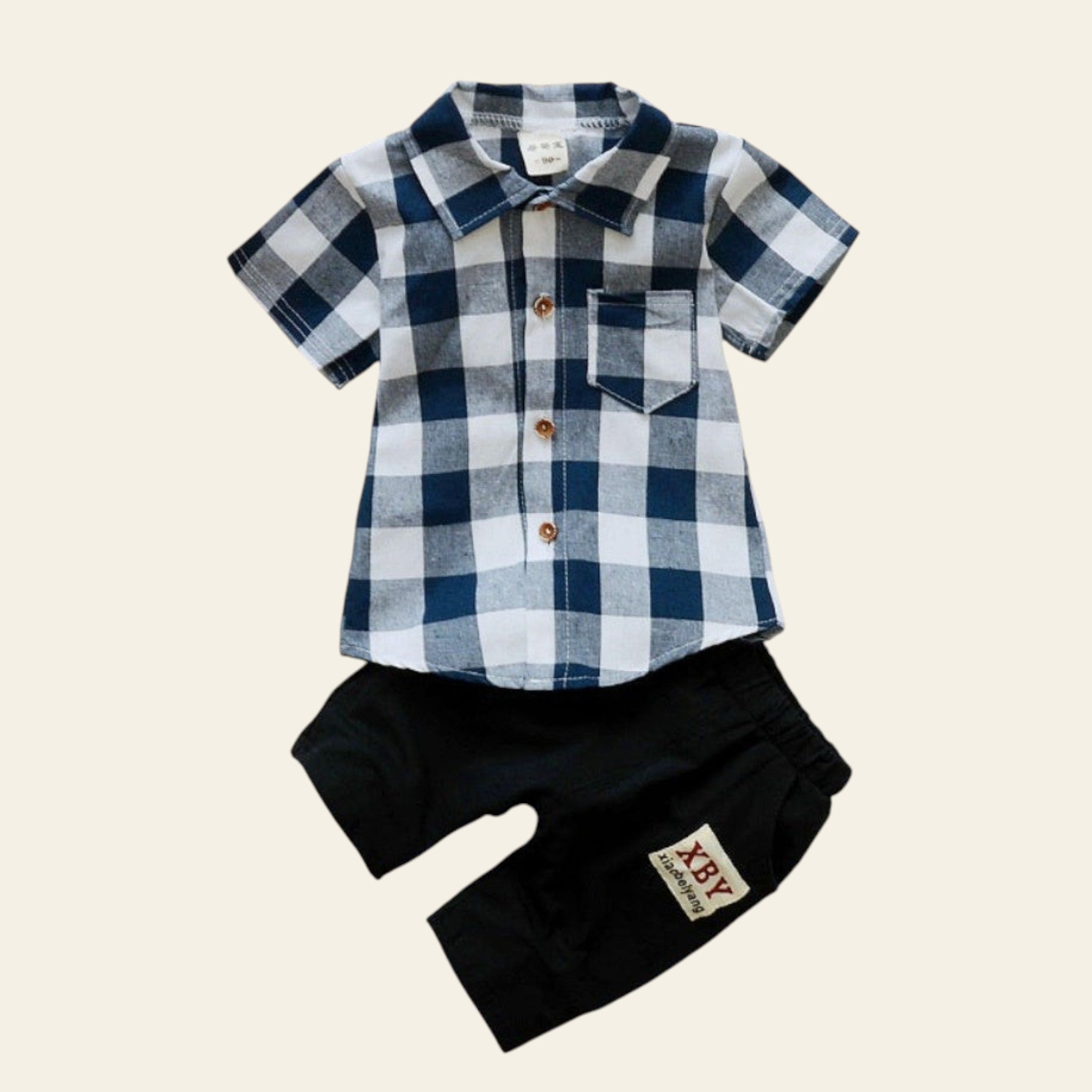 Children's plaid shirt and black shorts set on a wooden surface with flowers and pinecones.