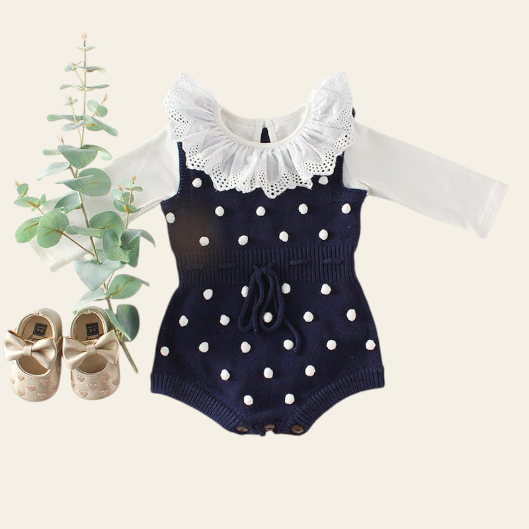 Navy blue romper with white polka dots and lace collar on a light gray background with baby shoes and greenery.