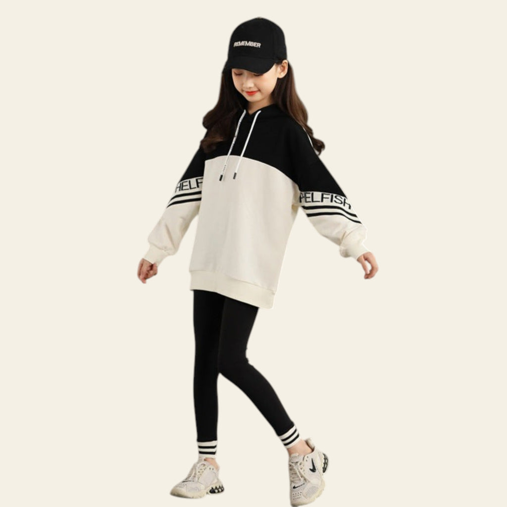 Child wearing a black and white hoodie with 'SELFISH' text, black cap, and black leggings against a gray concrete wall.