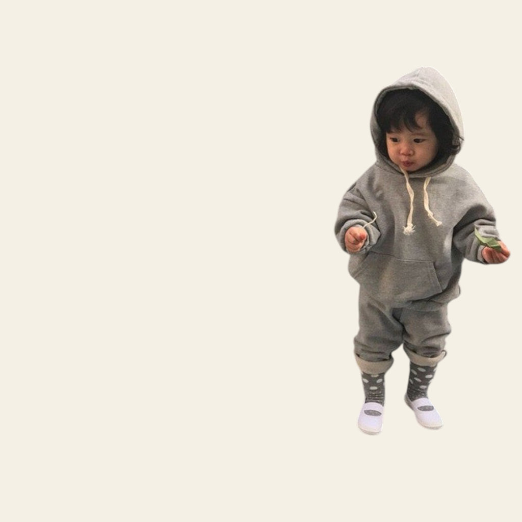 Child wearing a gray hoodie and pants standing against a plain wall.