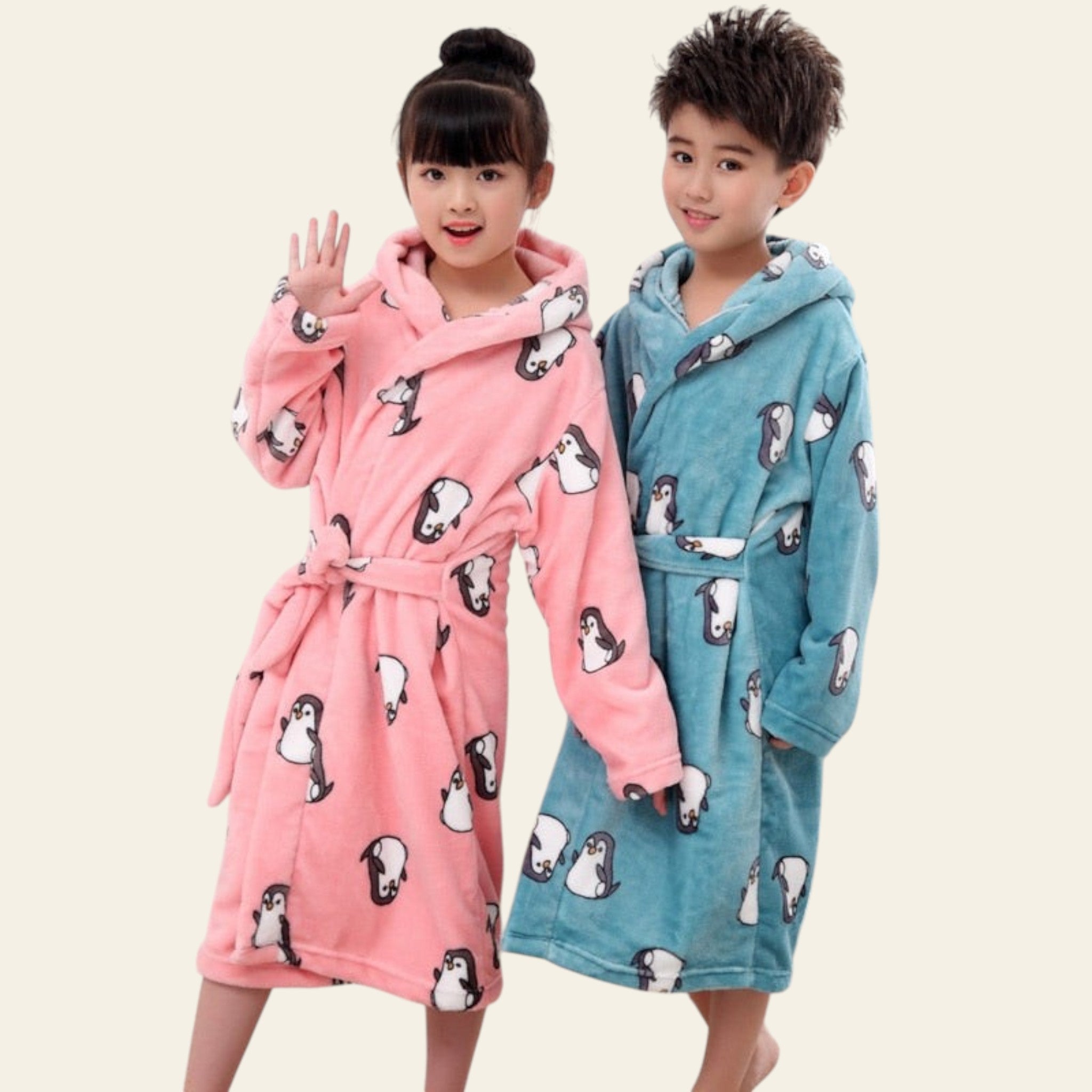 Two children wearing pink and blue hooded robes with penguin patterns in a living room setting.
