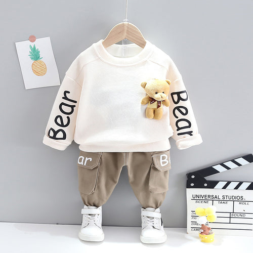 Children's outfit with white shirt and beige pants on a hanger against a gray wall.