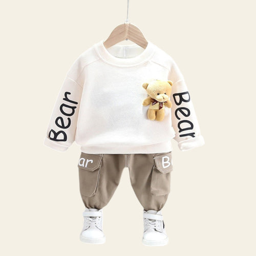 Children's outfit with white shirt and beige pants on a hanger against a gray wall.