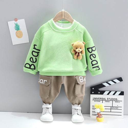 Children's green sweatshirt with bear design and brown pants on a hanger against a gray wall.