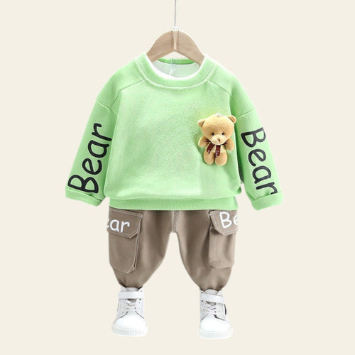 Children's green sweatshirt with bear design and brown pants on a hanger against a gray wall.