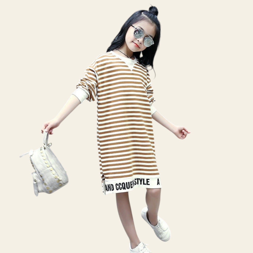 Young girl wearing a striped dress with visible branding, standing outdoors.