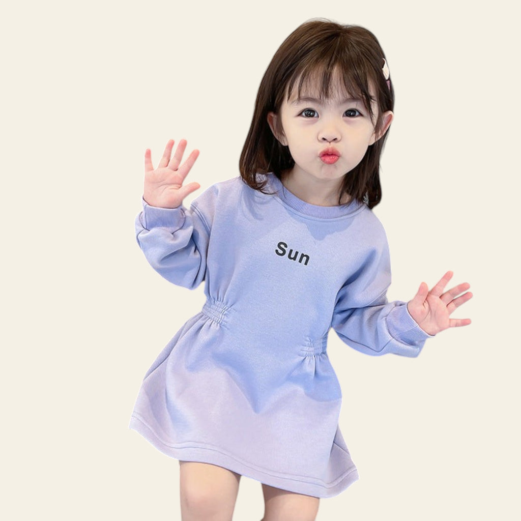 Child in a light blue dress with 'Sun' printed on it, standing in a kitchen.