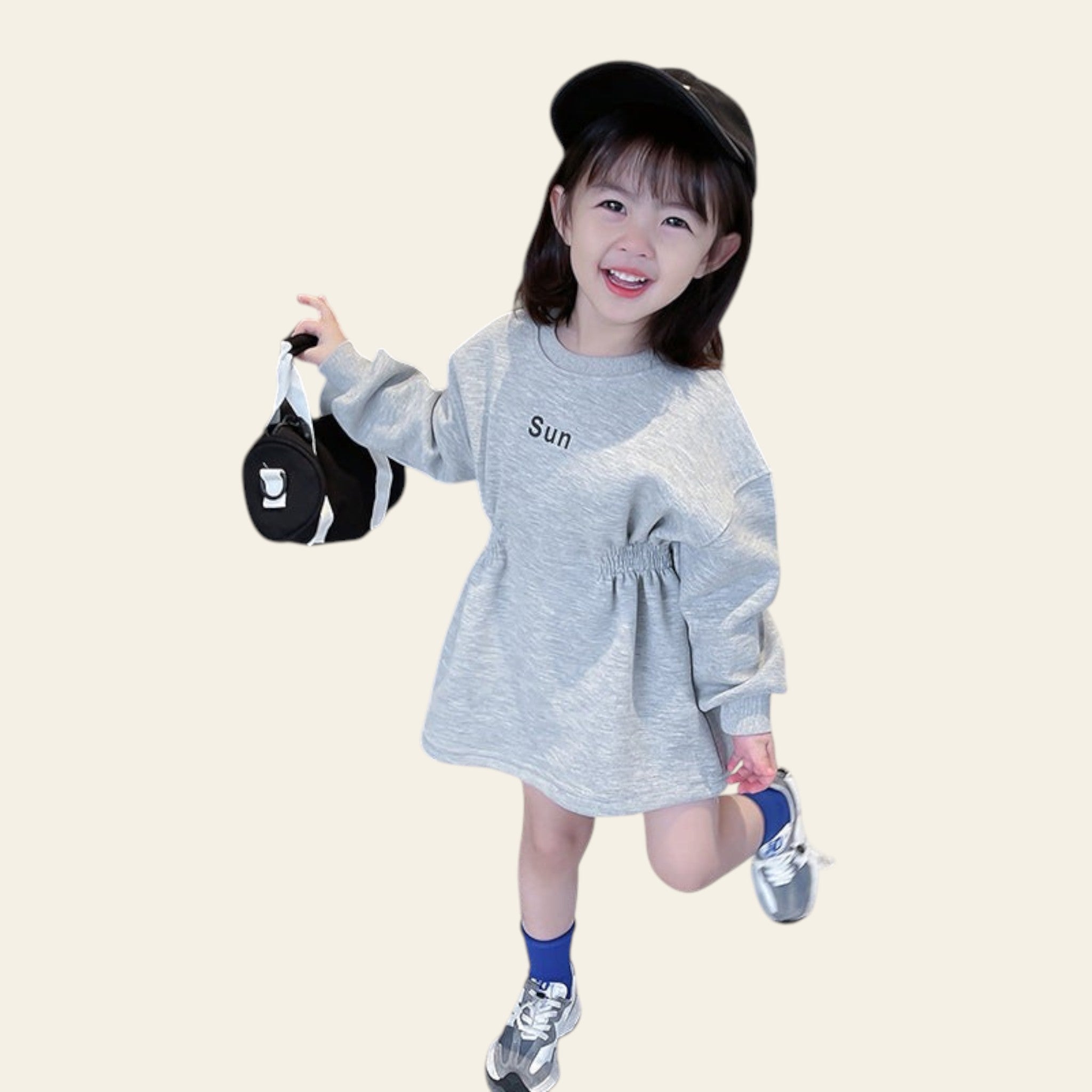 Child in a gray dress with 'Sun' printed on it, holding a black and white bag, standing on a concrete surface.