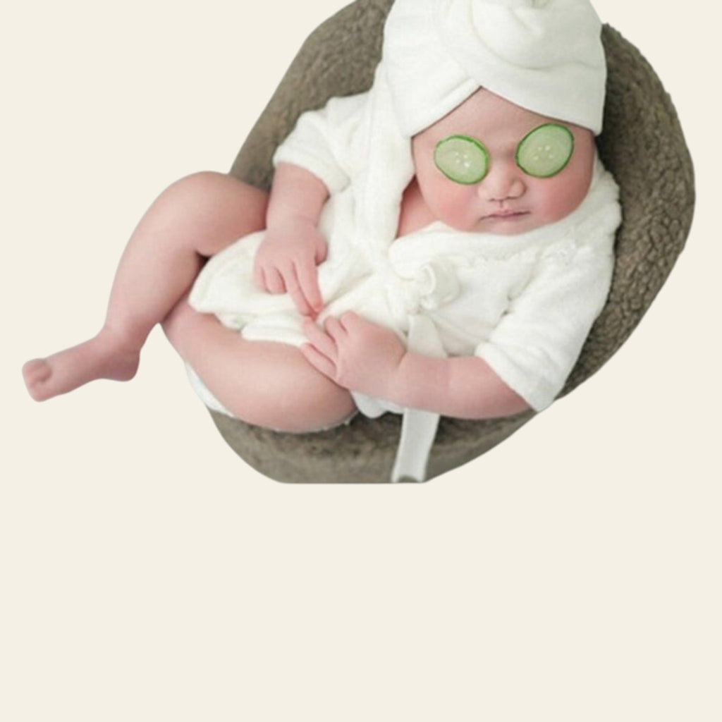 Baby in a white robe with cucumber slices on eyes, surrounded by baby care items.