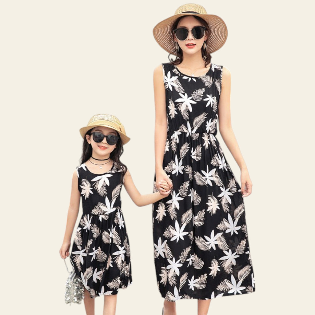 Two people wearing matching black floral dresses with sunglasses and hats, standing by a poolside.
