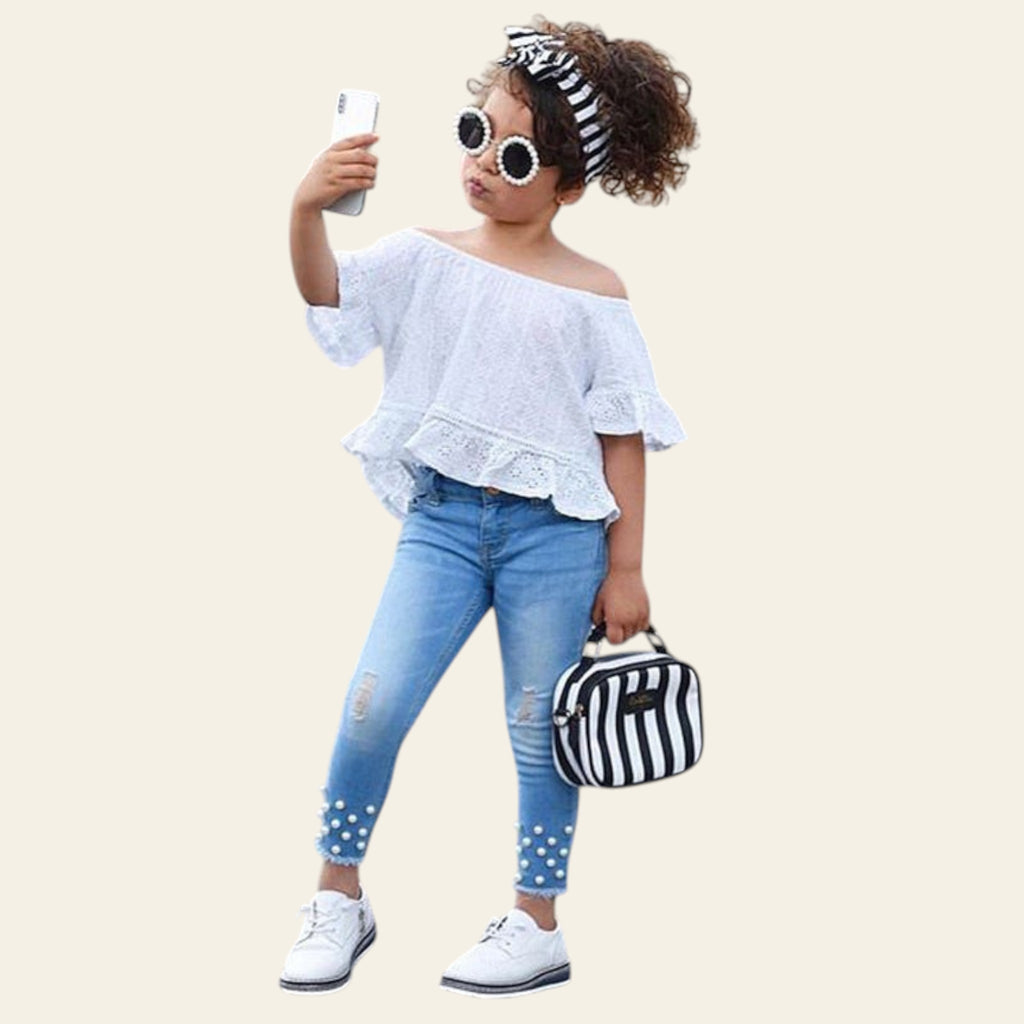 Child in white off-shoulder top and blue jeans holding a phone on a street.