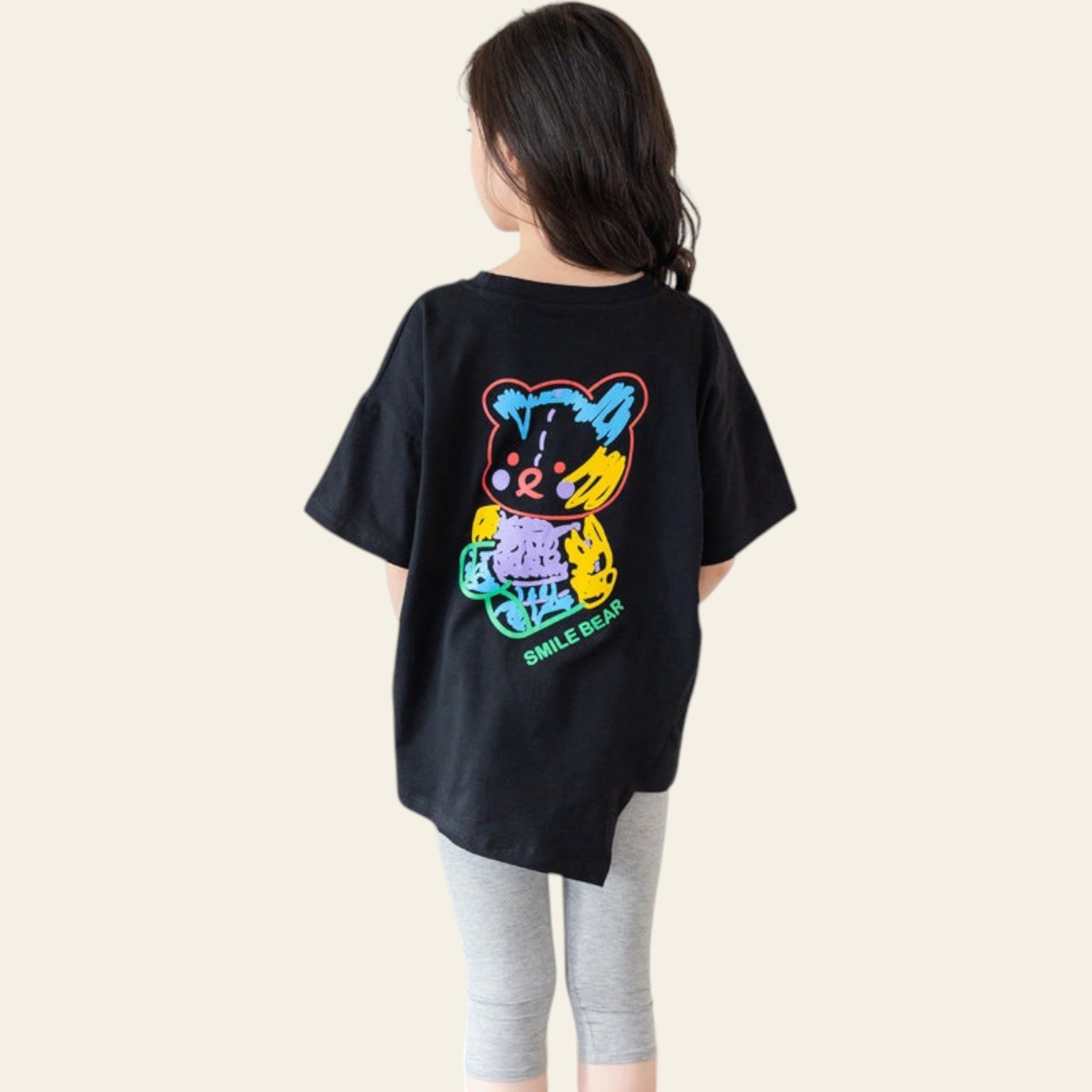 Person wearing a black t-shirt with a colorful bear graphic on the back, standing in a room with a wooden chair and white wall.
