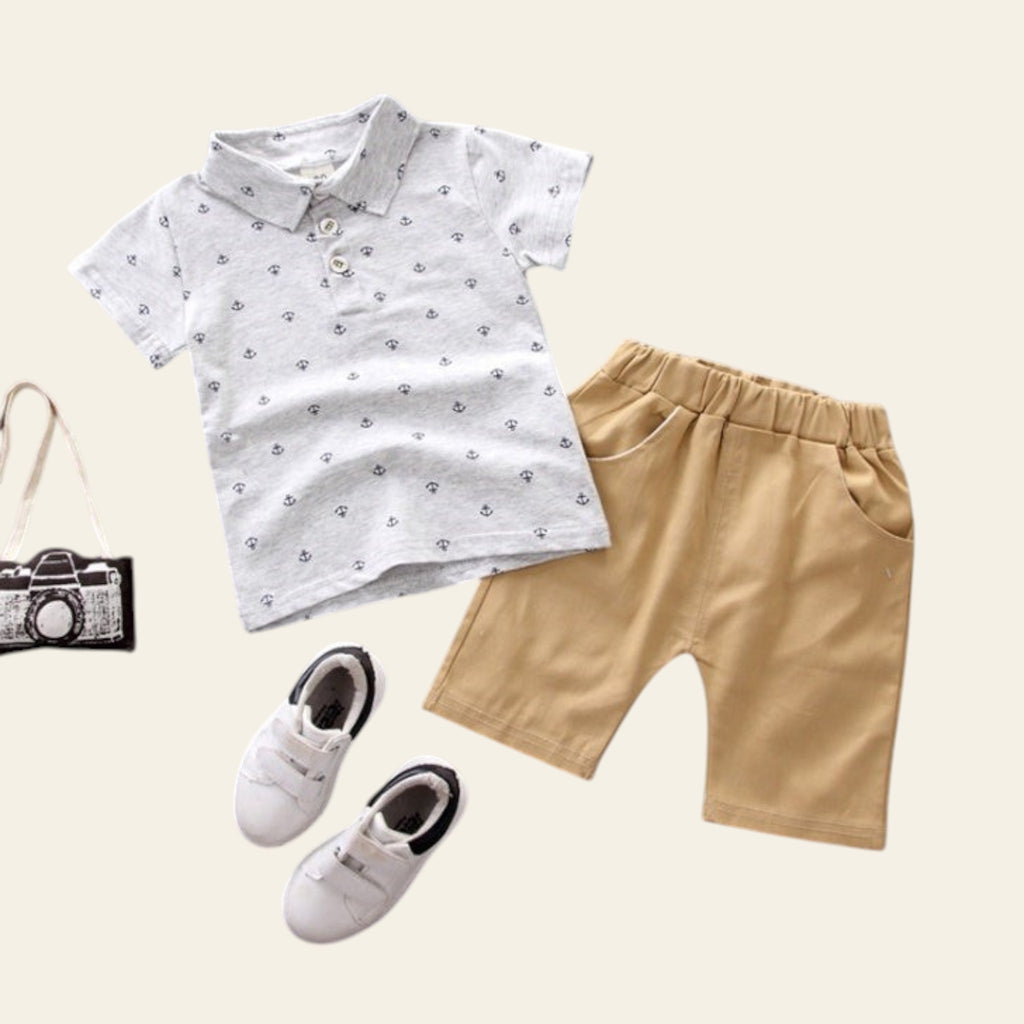 Children's outfit with white patterned shirt, beige shorts, white shoes, and a small black camera bag on a gray background.