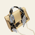 Beige handbag with a patterned scarf and gold chain strap on a white surface.
