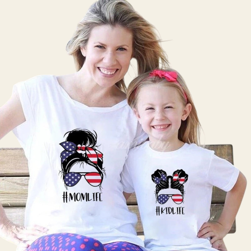 Woman and child wearing matching white t-shirts with 'Momlife' and 'Kidlife' designs, sitting outdoors.