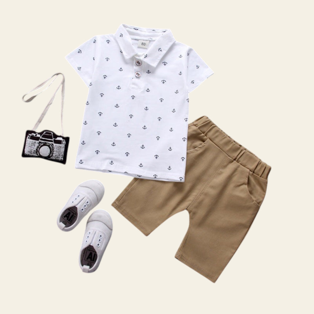 Children's outfit with white polo shirt, brown shorts, white sneakers, and small camera bag on a gray background