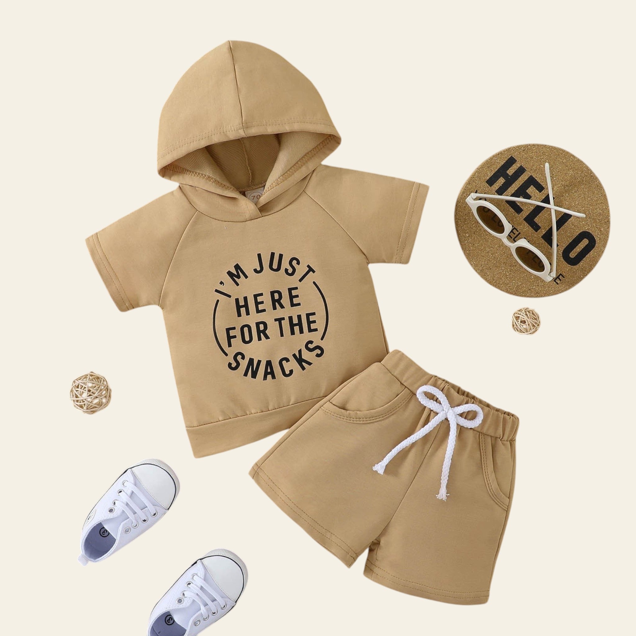 Beige hoodie and shorts set with 'I'm just here for the snacks' text on a wooden surface, accompanied by white sneakers and decorative items.