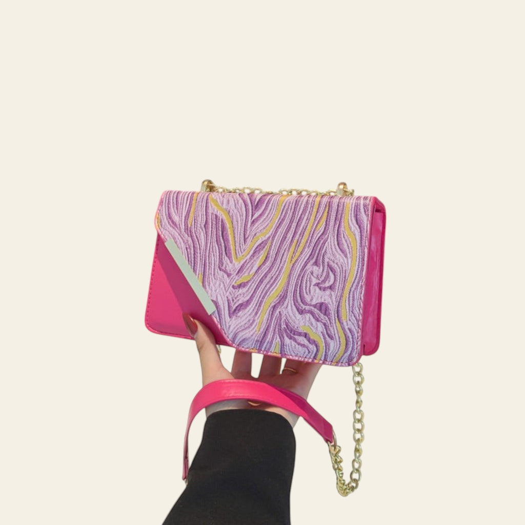 Hand holding a pink and purple patterned handbag with a chain strap against a black background.