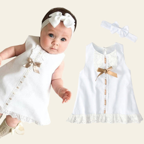 Baby wearing a white outfit with lace details and a matching headband on a textured white surface.