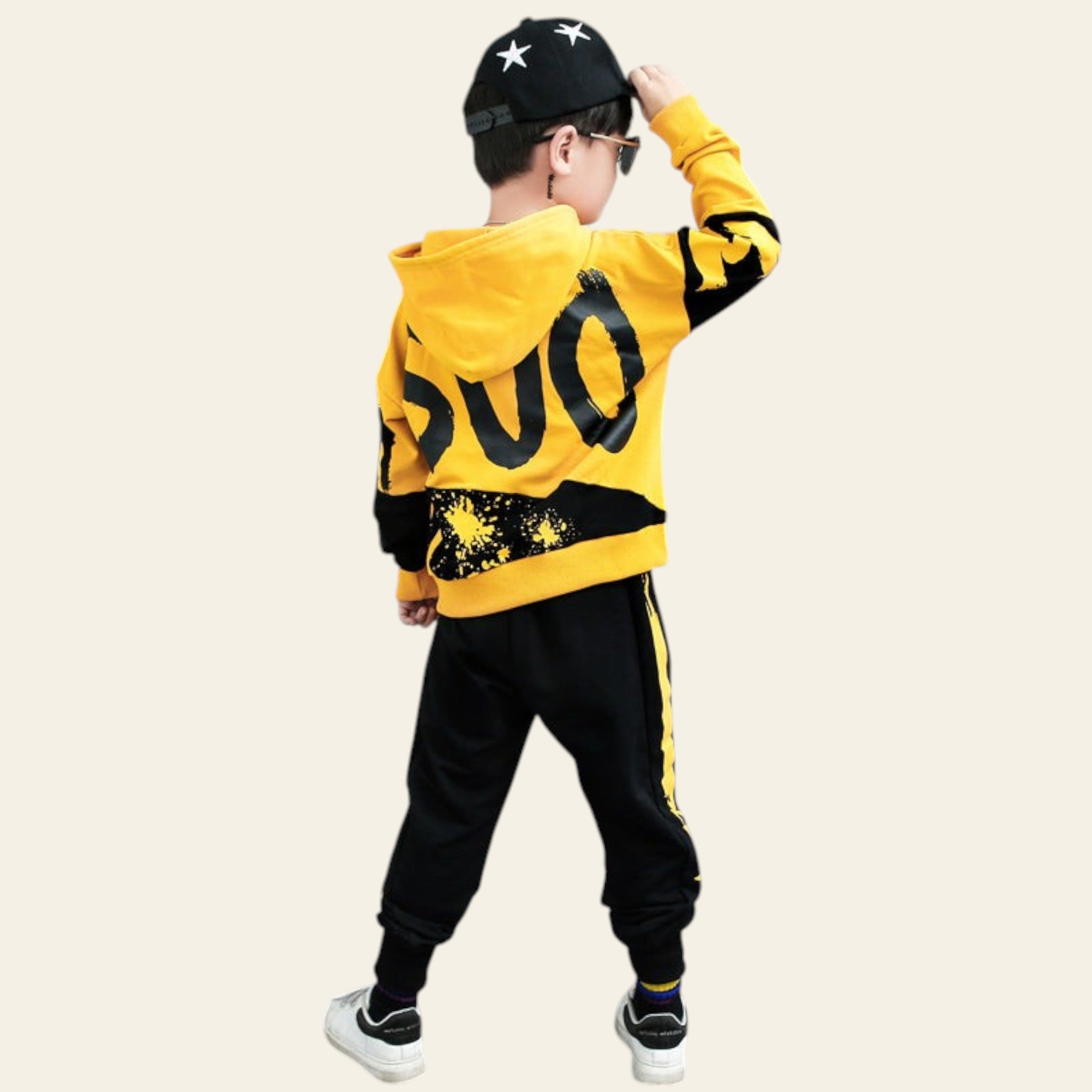 Child wearing a yellow hoodie with black lettering and black pants against a gray background with large letters.