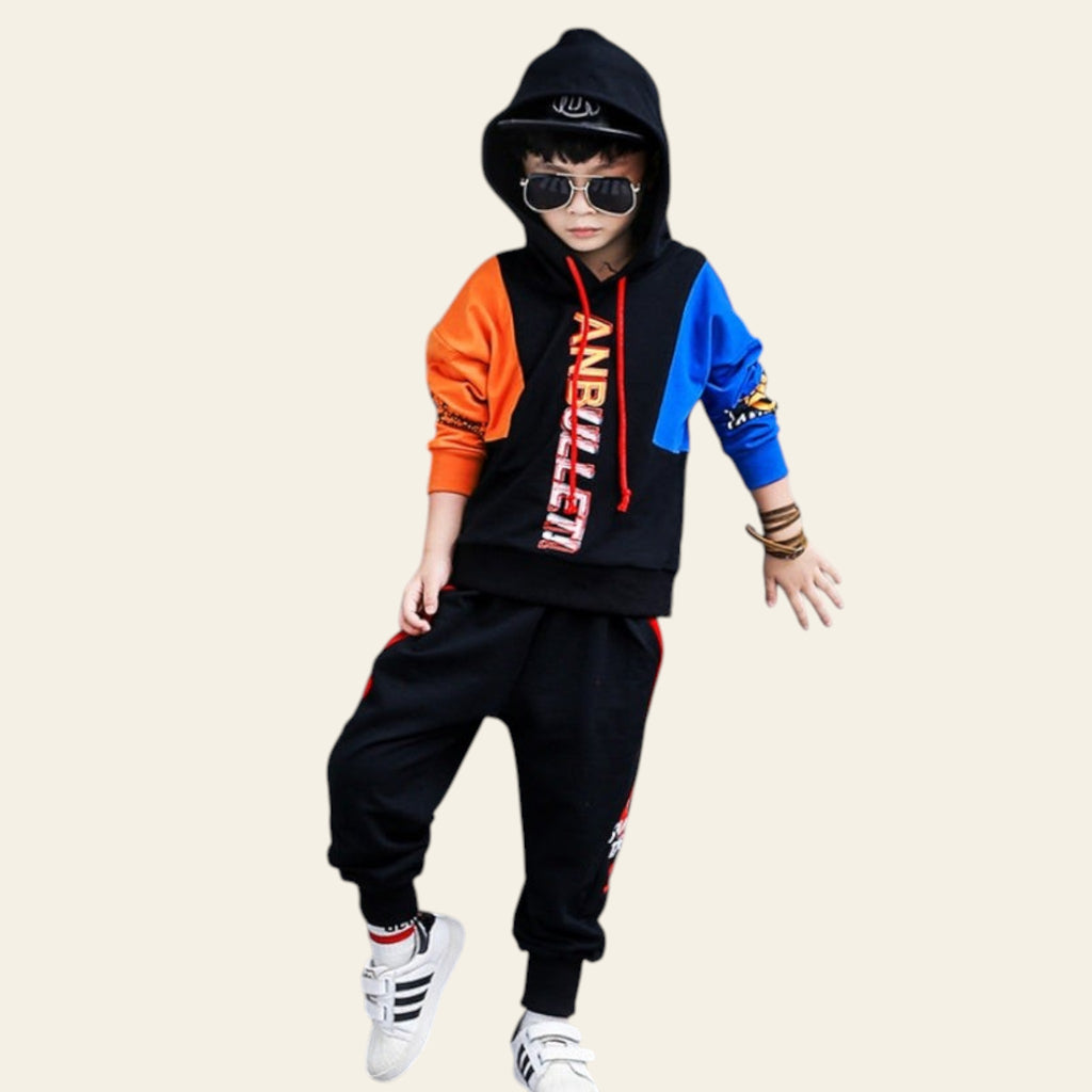 Child wearing a colorful outfit with a black hoodie and pants, standing against a gray wall with large black letters.