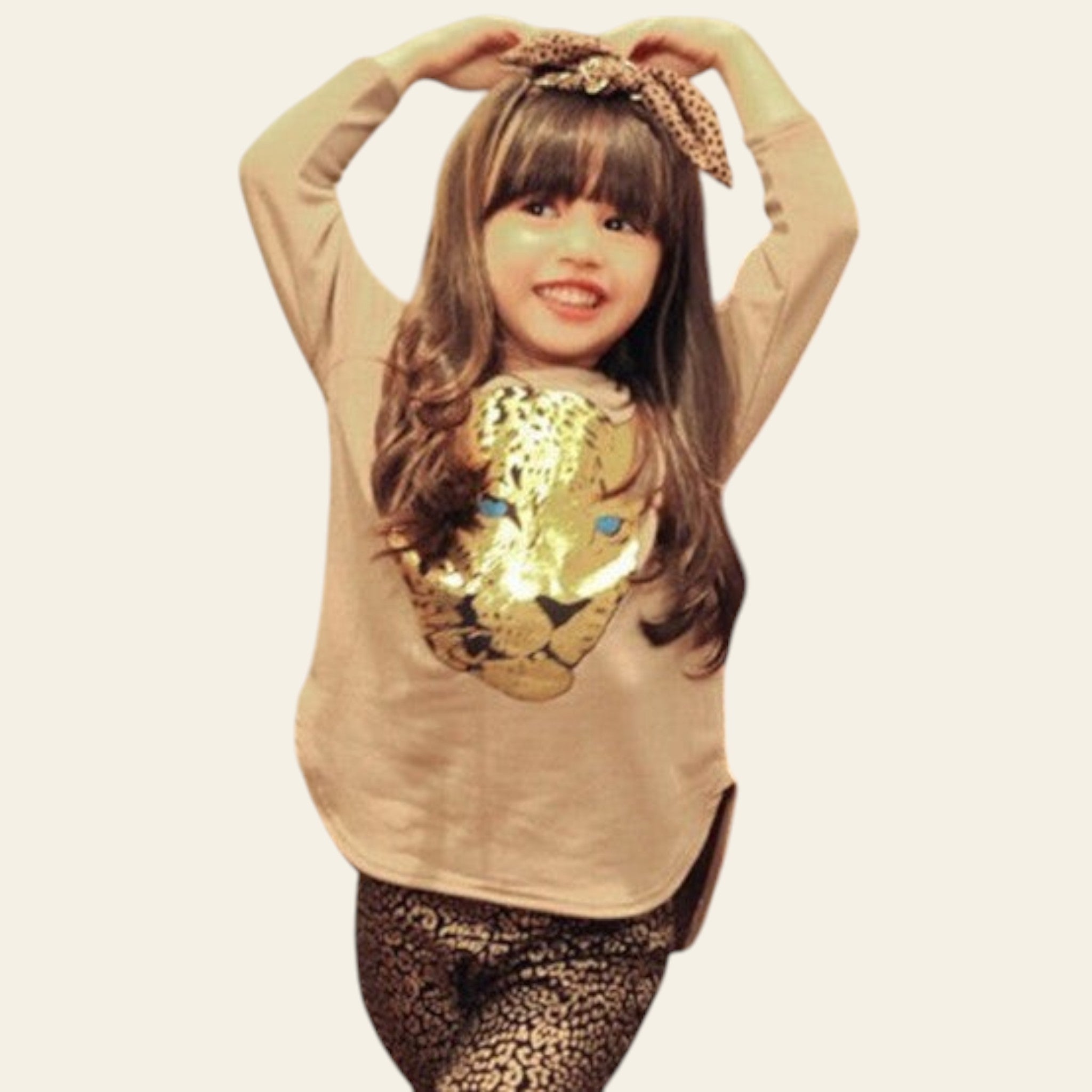 Young girl in a beige sweater with a sequin design and leopard print pants posing indoors.