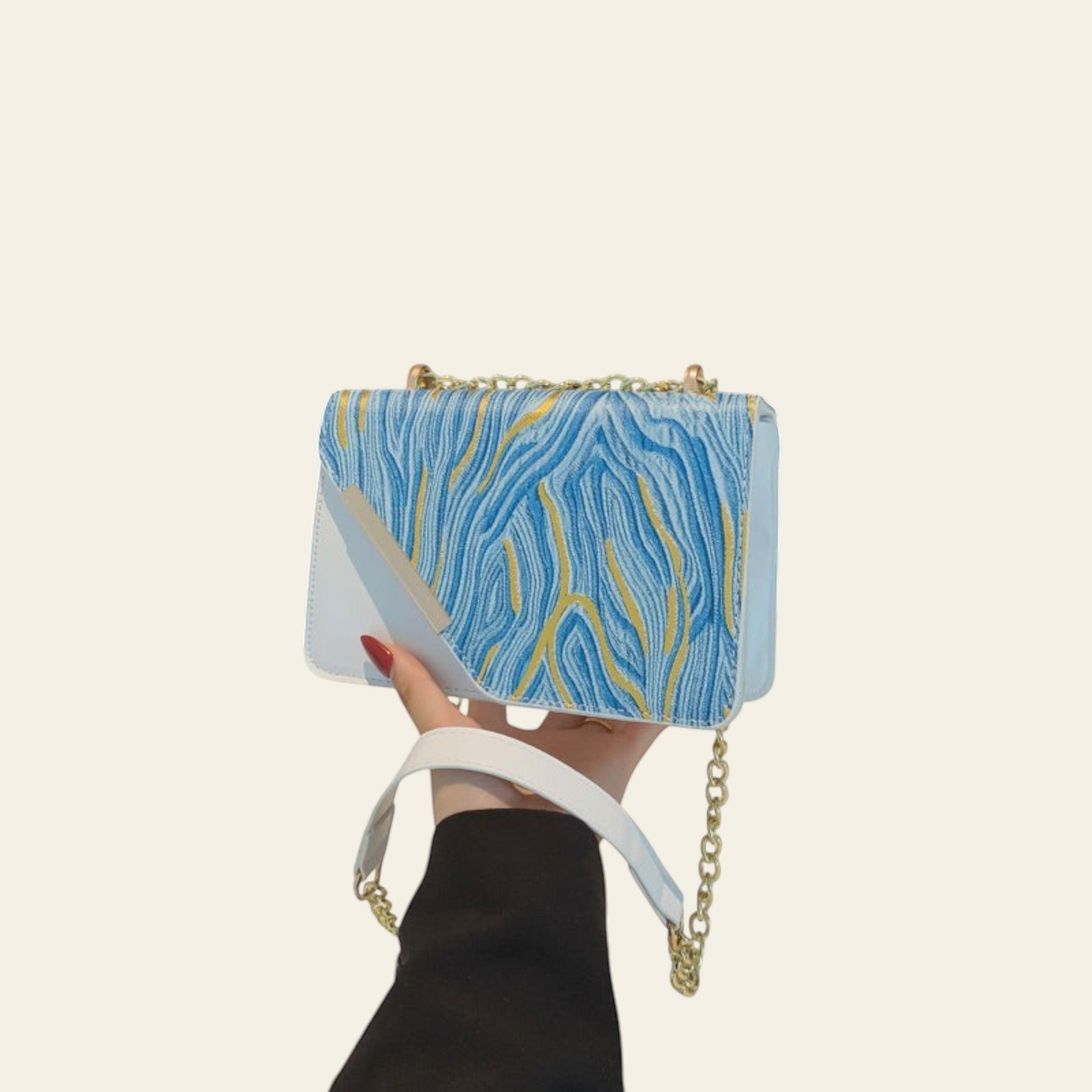 Hand holding a blue and gold marbled handbag with a chain strap against a black background.