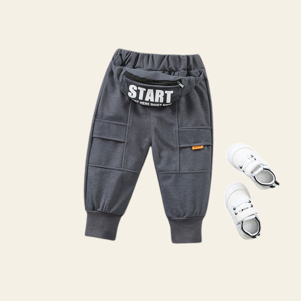 Gray children's jogger pants with 'START' text and white sneakers on a textured gray surface.