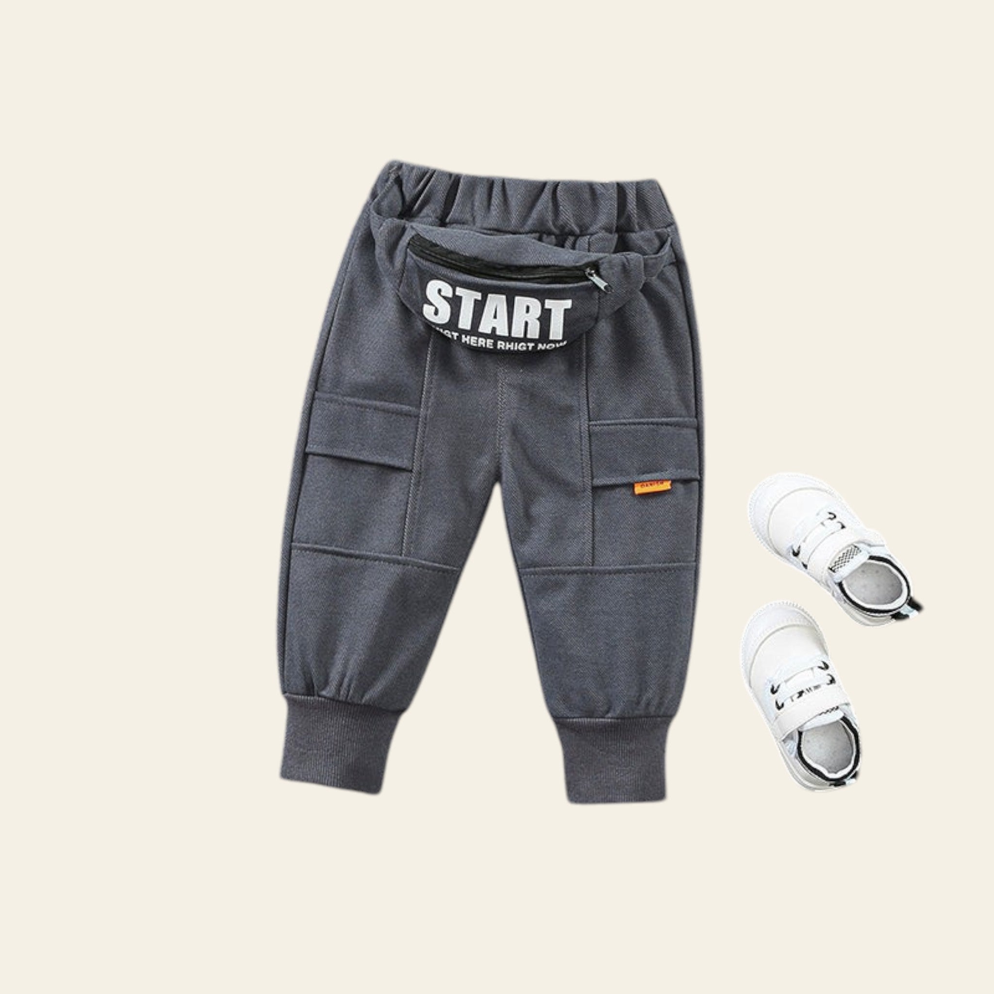 Gray children's jogger pants with 'START' text and white sneakers on a textured gray surface.