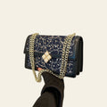 Black handbag with gold chain and embellishments held by a hand against a white wall.