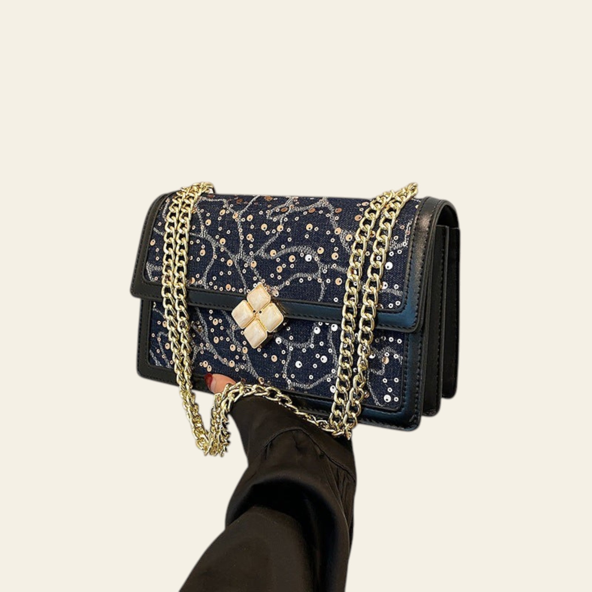 Black handbag with gold chain and embellishments held by a hand against a white wall.