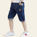Child wearing blue denim shorts with a logo, standing on a wooden floor against a gray wall.