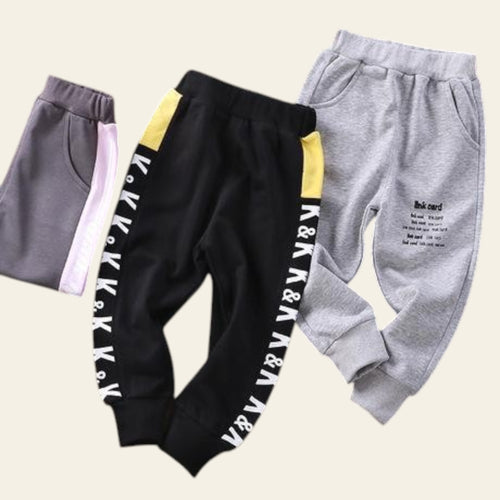 Three pairs of children's sweatpants in gray, black, and gray with text on a wooden surface.