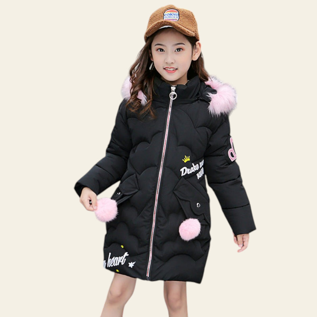Child wearing a black puffer coat with pink pom-poms and a brown cap, standing on a street.