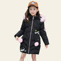 Child wearing a black puffer coat with pink pom-poms and a brown cap, standing on a street.