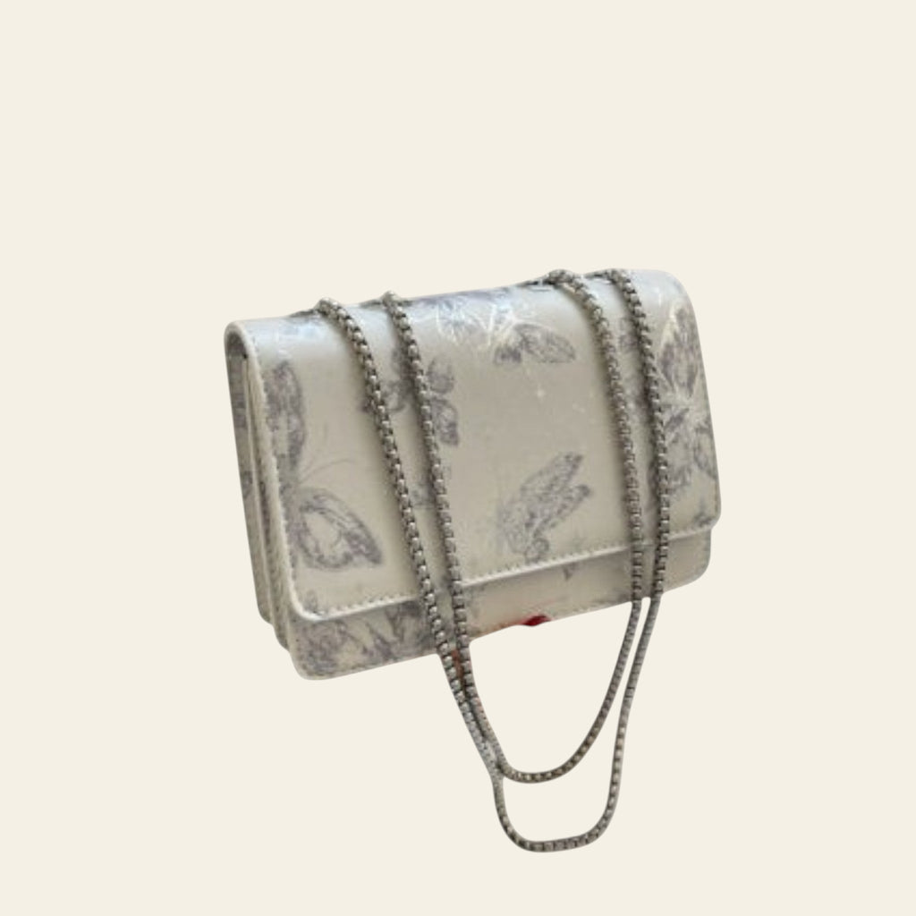 Silver floral handbag with chain detail held by a person, with a gray coat and glass of water in the background.