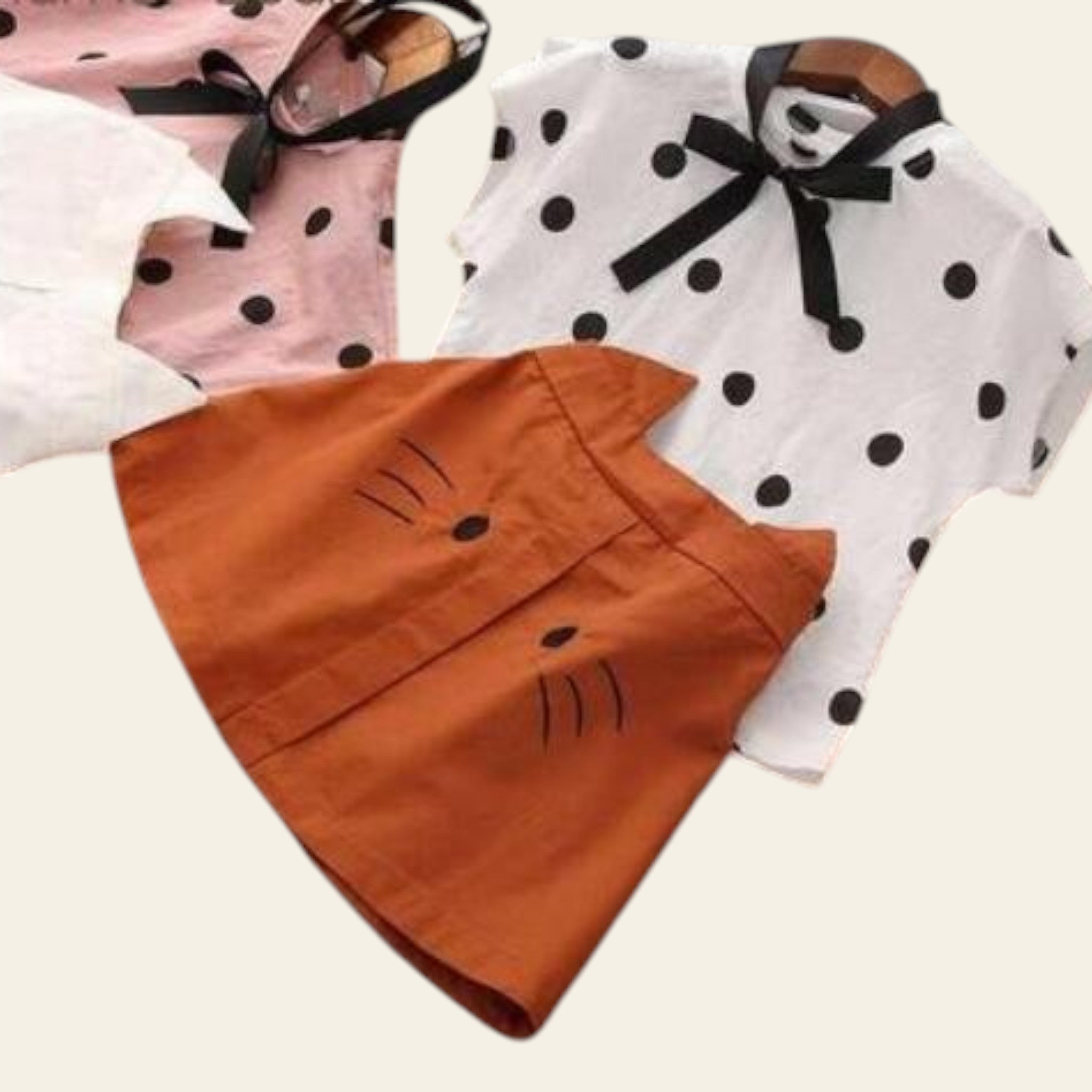 Three children's hats on a wooden surface, including a pink one with black polka dots, a white one with black polka dots, and an orange one with cat face design.