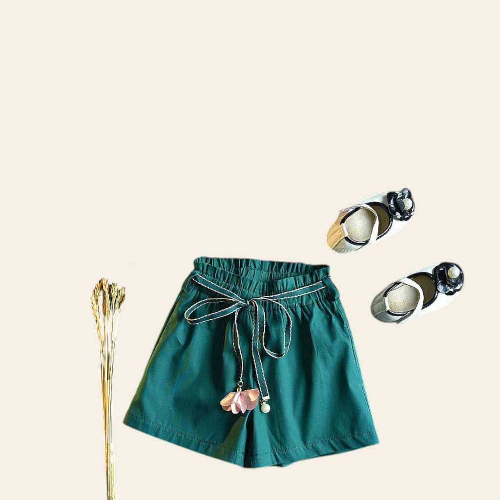 Green shorts with decorative elements, straw hat, sunglasses, and dried plants on a textured surface