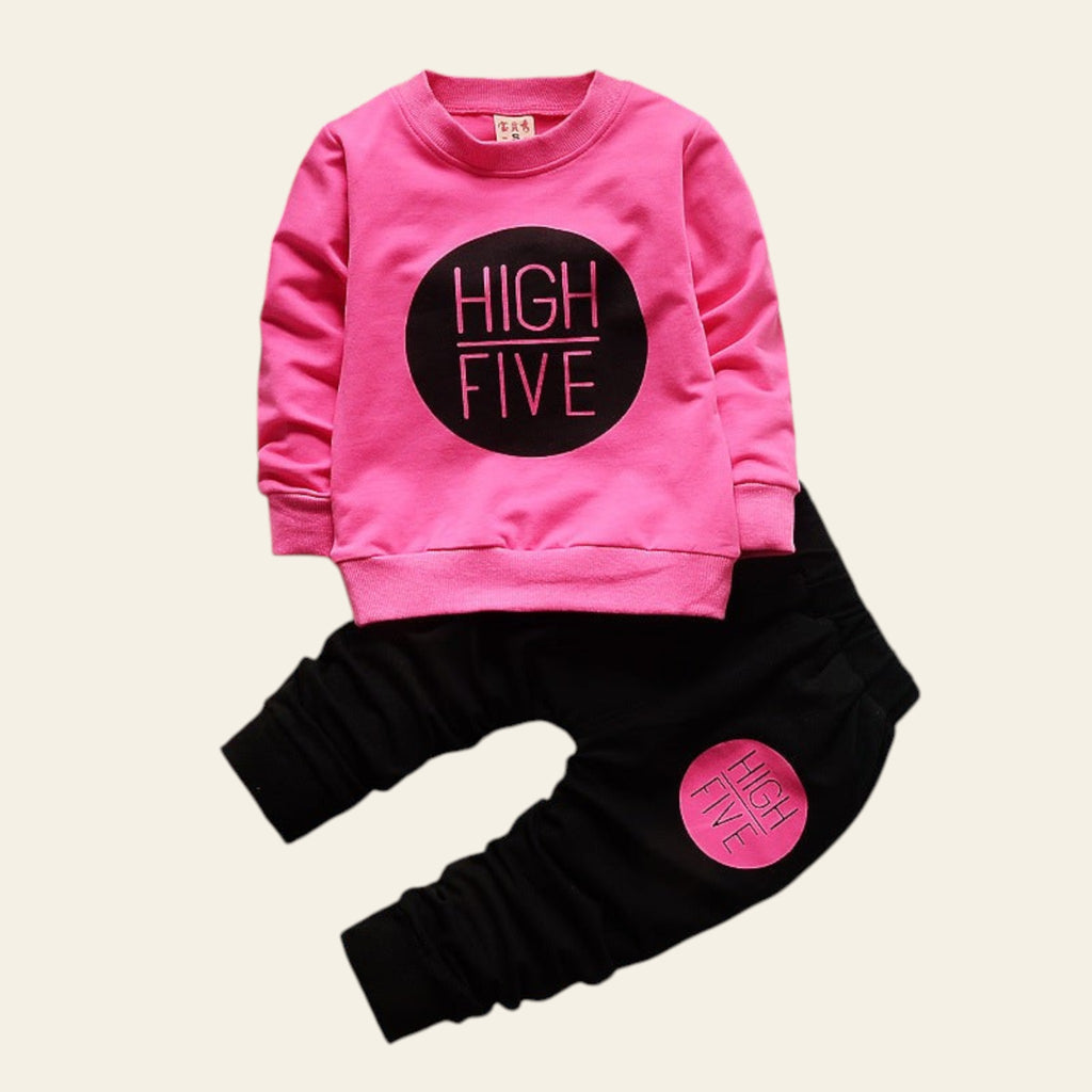 Pink sweatshirt and black pants set with 'HIGH FIVE' text on a wooden surface with decorative elements.