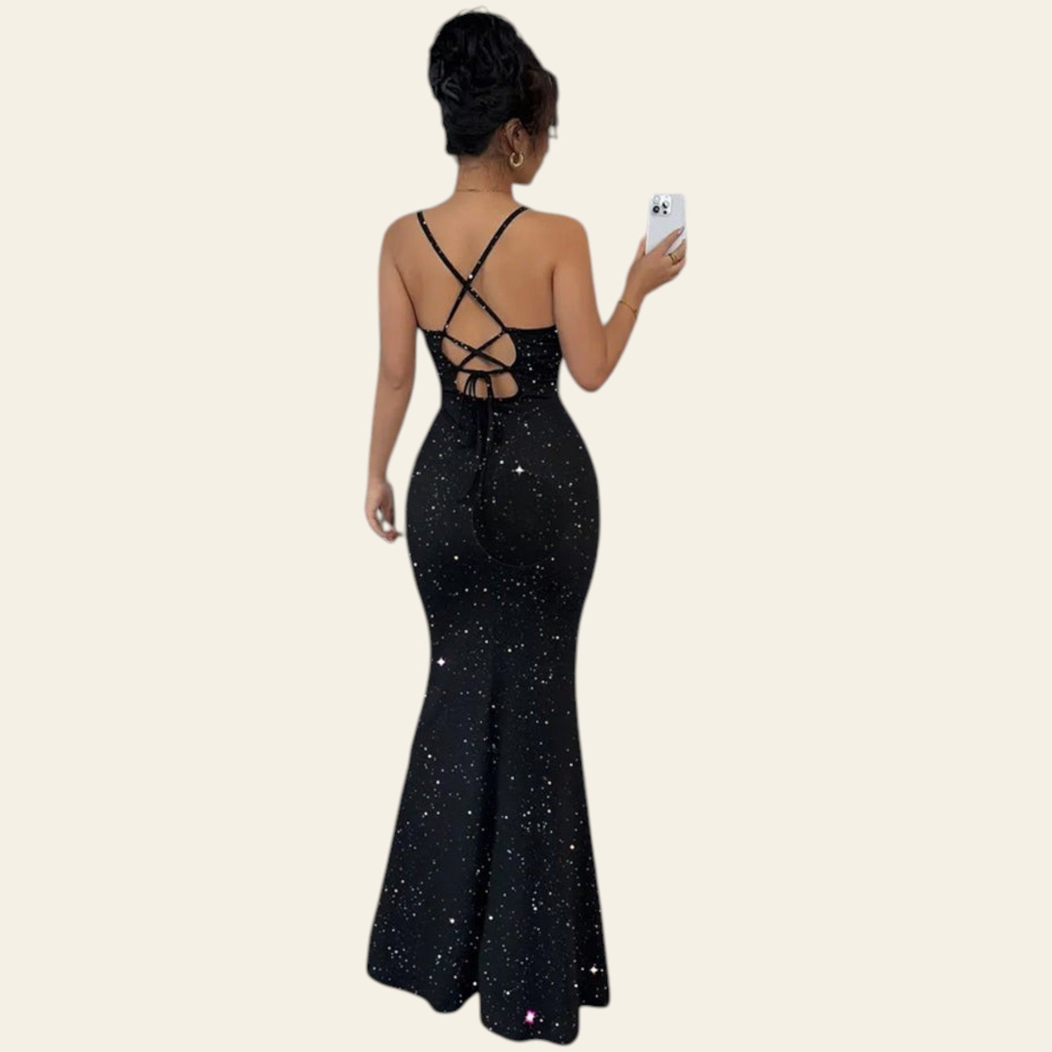 Back view of a woman wearing a black backless sequin maxi slip dress with crisscross straps, standing in front of a silver party backdrop