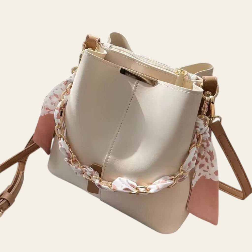 Beige handbag with a decorative chain and scarf on a mannequin, placed on a surface.