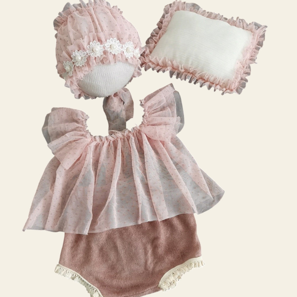 Baby outfit with pink top and bloomers, white headband, and pink bonnet on a wooden surface.