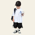 Child wearing a white t-shirt, black shorts, and a cap, holding a drink on a wooden floor.