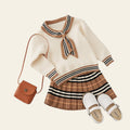 Children's outfit with sweater, skirt, shoes, and handbag on a textured surface.